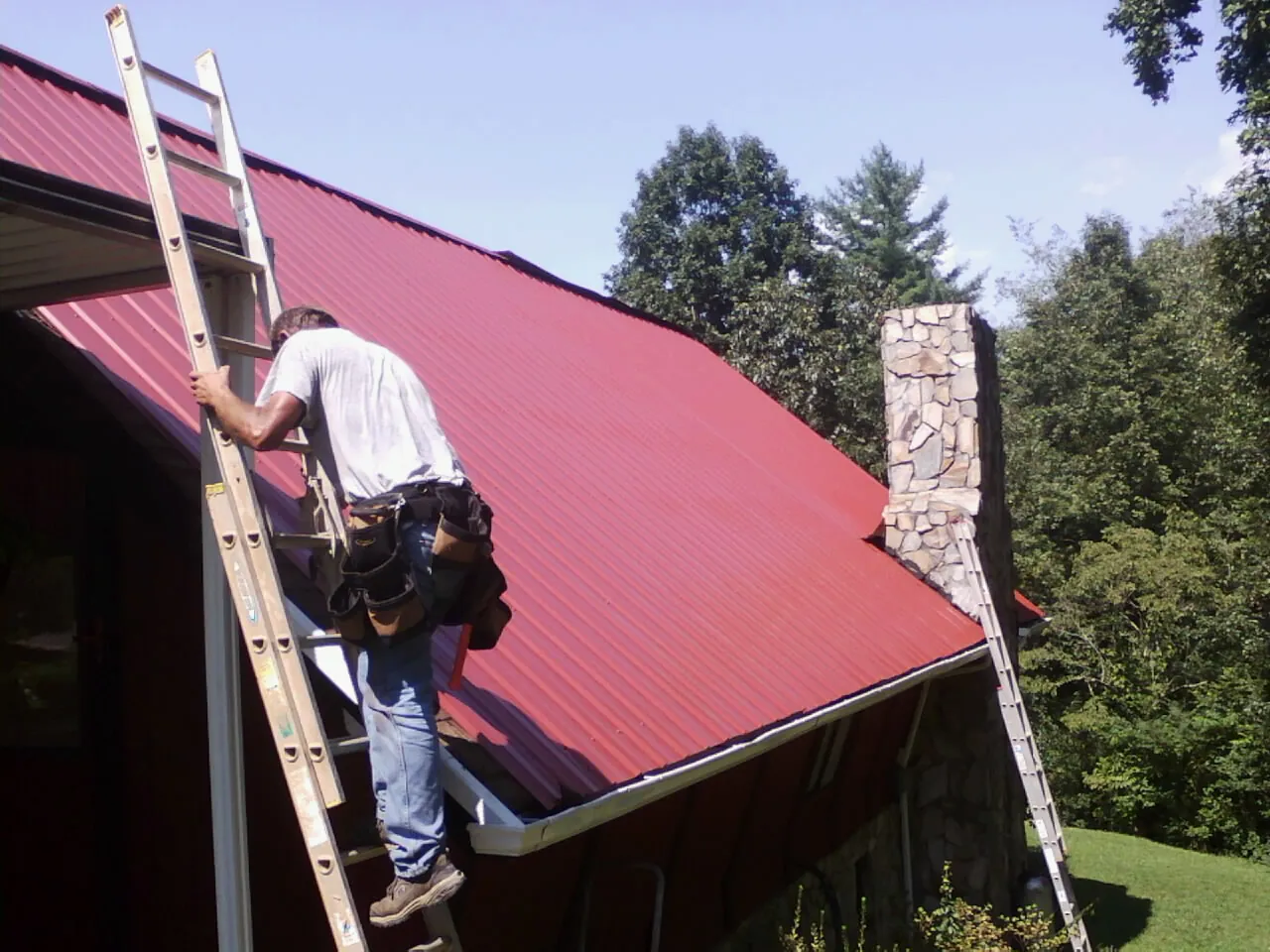 Roofer on a ladder beside a red metal roof and stone chimney — Soffit & Fascia Repair by Roofing Brothers in Chimacum